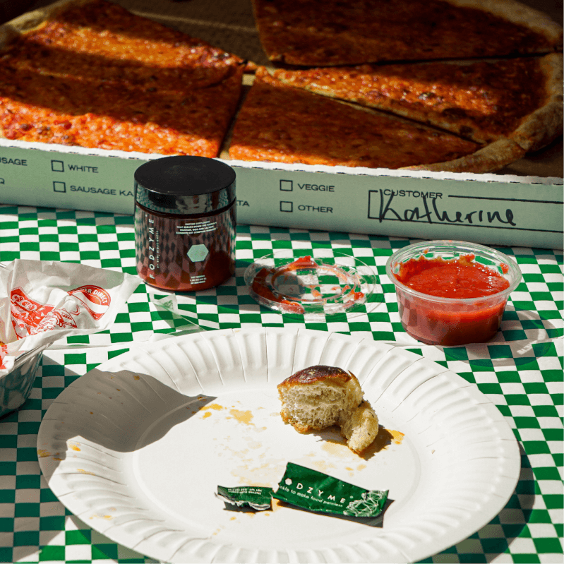 FODZYME enzyme powder jar and single-serve packet alongside a pizza box and tomato dipping sauce on a green checkered tablecloth.