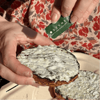 Hands holding a toasted bagel with cream cheese while opening a FODZYME single-serve packet to sprinkle on top.