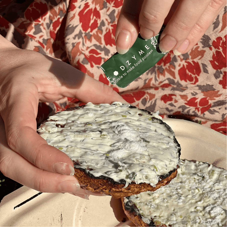 Hands holding a toasted bagel with cream cheese while opening a FODZYME single-serve packet to sprinkle on top.