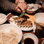 Hand holding a FODZYME packet over a plate of pan-fried dumplings at a restaurant table.
