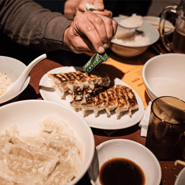 Hand holding a FODZYME packet over a plate of pan-fried dumplings at a restaurant table.